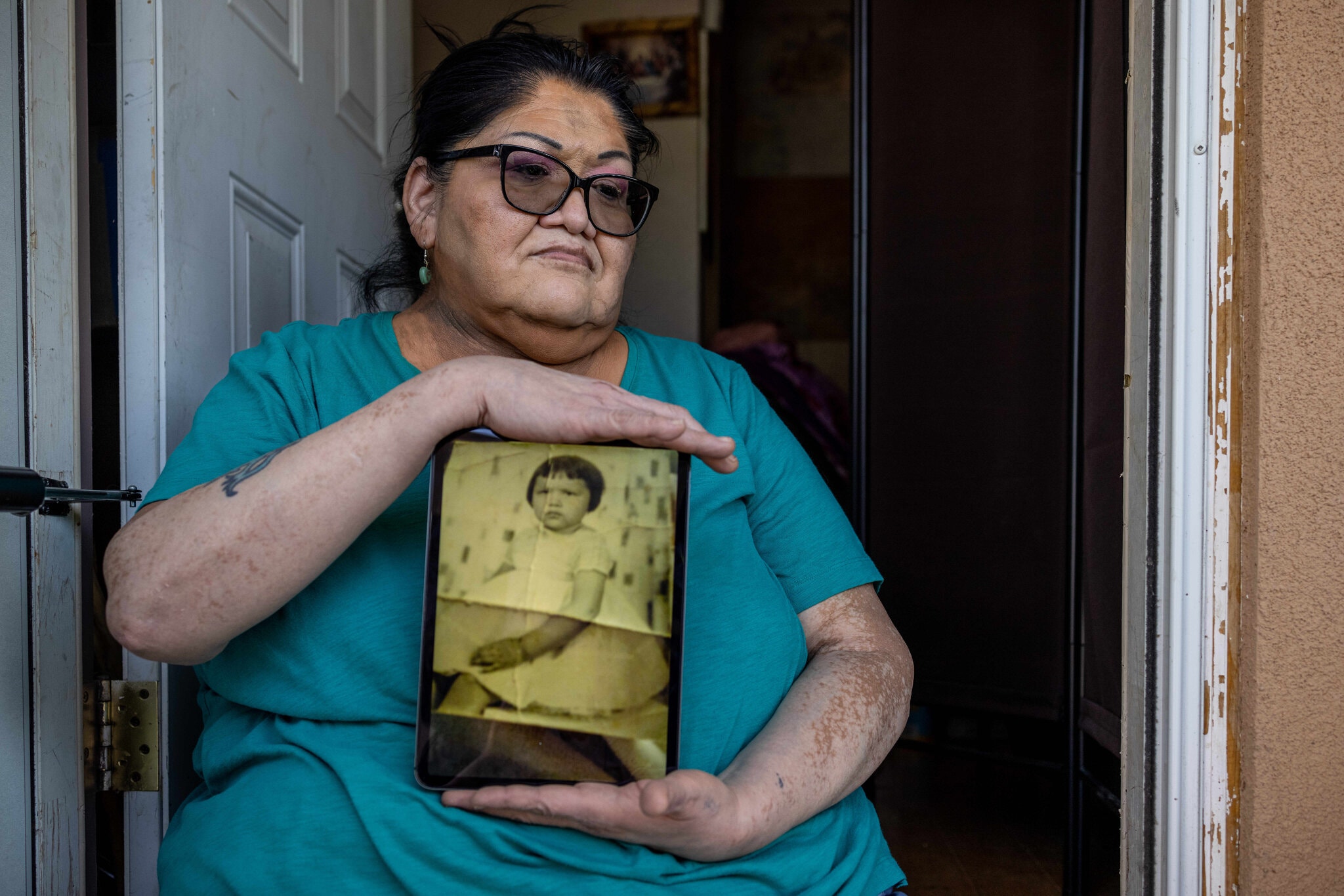 [Jacqueline Frost, 60, holds a photo showing how she was forced to adopt the look and attire of a white girl. She said she was beaten by a Ute aunt who served as a matron at a federal boarding school designed to assimilate Native children.Credit...Sharon Chischilly for The New York Times]